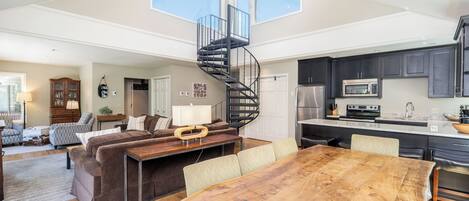 A spacious living area with a wooden dining table, a sofa set, a spiral staircase, and a kitchen with dark cabinets and modern appliances.