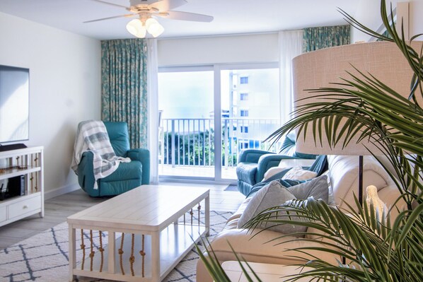 Un salon moderne avec un canapé, des fauteuils, une table basse et un téléviseur. Il y a un balcon avec vue sur les immebles.