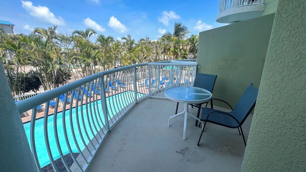 Un balcon doté d’une table et de chaises en verre, offrant une vue sur une piscine et des palmiers.