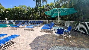 Un espace aménagé au bord de la piscine, avec des chaises longues bleues et une table, le tout entouré d’une clôture blanche et de palmiers.