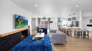 Un salon moderne avec une cheminée, des canapés bleus et gris, un coin repas avec une cuisine en arrière-plan et un grand tableau accroché au mur.