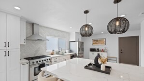Une cuisine moderne avec des meubles blancs, un grand îlot en marbre, des appareils en acier inoxydable et deux luminaires suspendus.