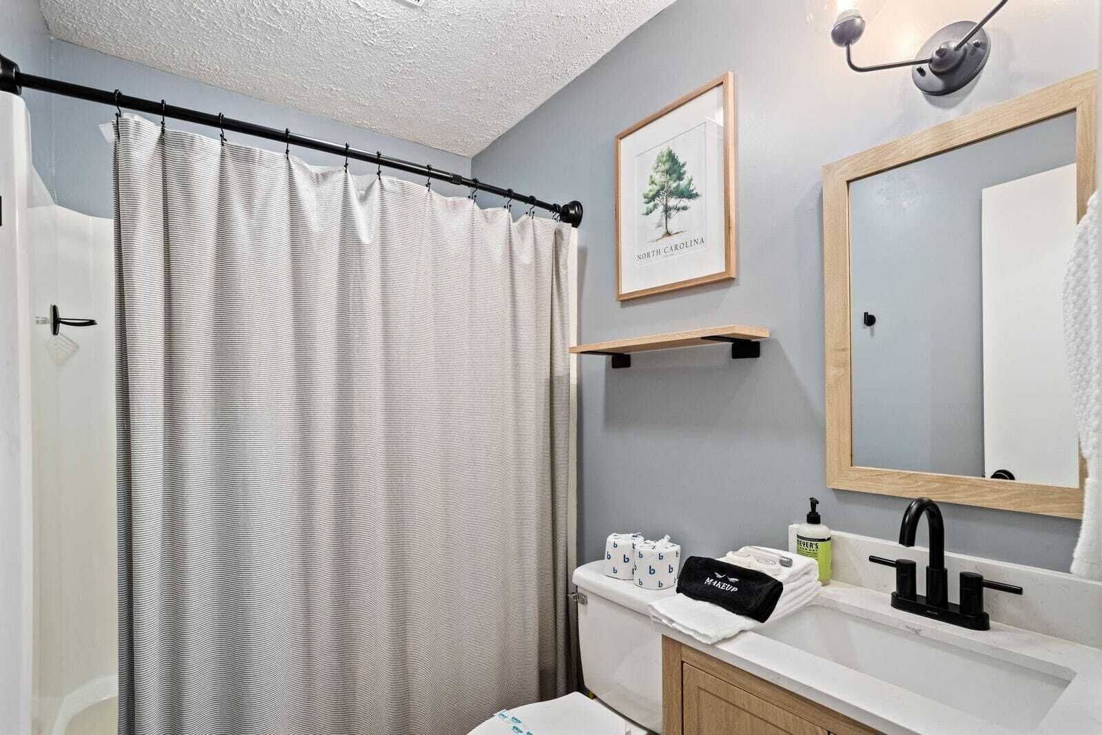 A bathroom with a striped shower curtain, a framed picture of a tree, a sink with toiletries, and a mirror.