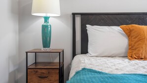 Una cama bien hecha con almohadas blancas y anaranjadas, una lámpara turquesa en una mesita de noche de madera y una pared blanca.