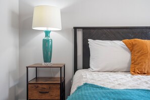 Una cama bien hecha con un almohadón blanco y un plaid naranja, una lámpara verde azulada sobre una mesita de noche de madera y un cajón debajo.