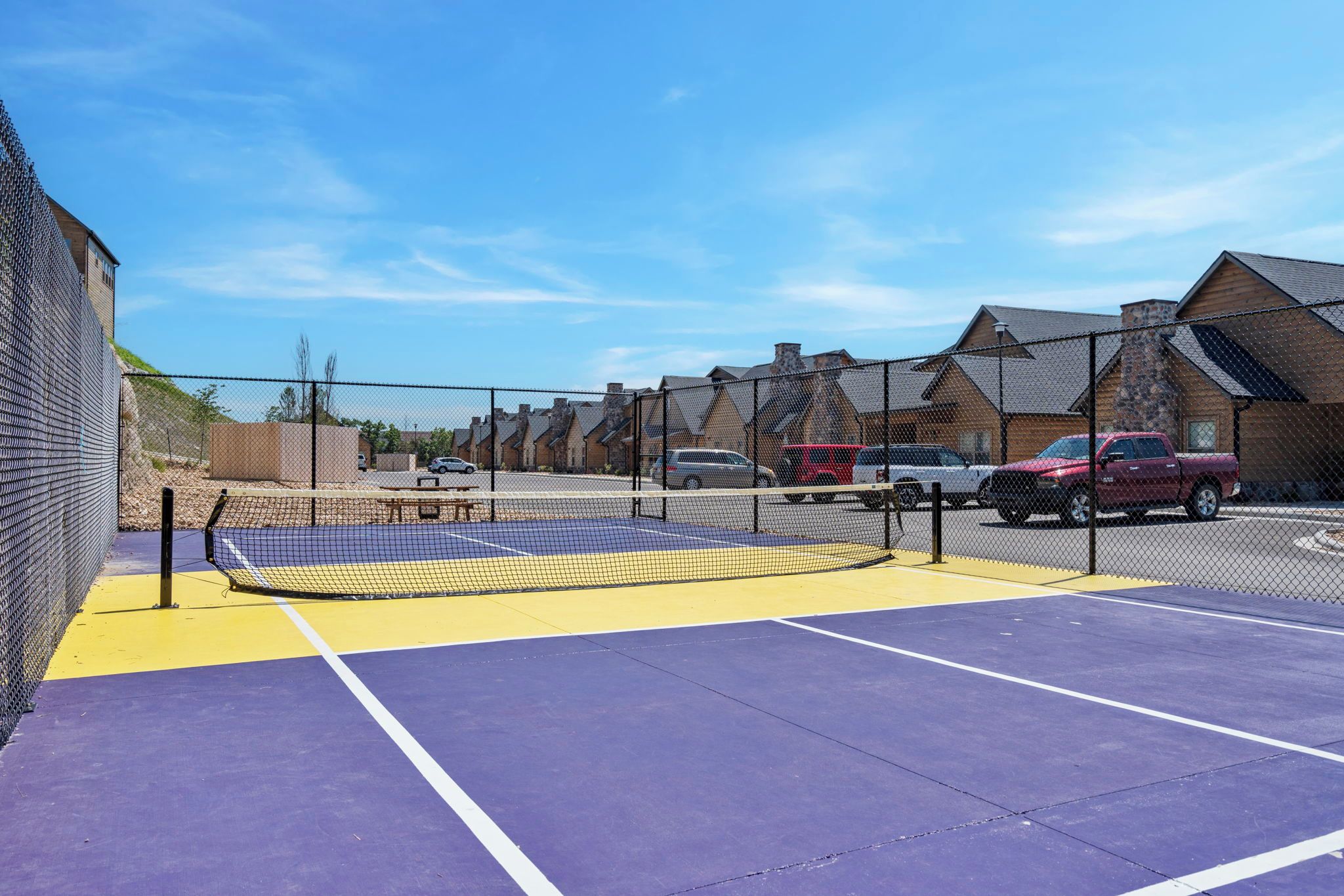 Un court de tennis avec un filet, entouré d’une clôture, et des véhicules garés à proximité.