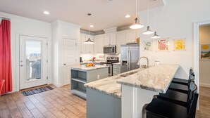 A modern kitchen with white cabinets, a large island, stainless steel appliances, pendant lighting, and a red curtain.