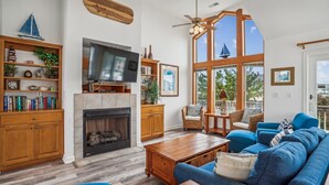 A spacious living room with a fireplace, a television, and a large window offering a view of the outdoors. - 1620 - Triple 7 - Corolla Sleeps 20 | Pool, Hot Tub & Beach Walk (Corolla)