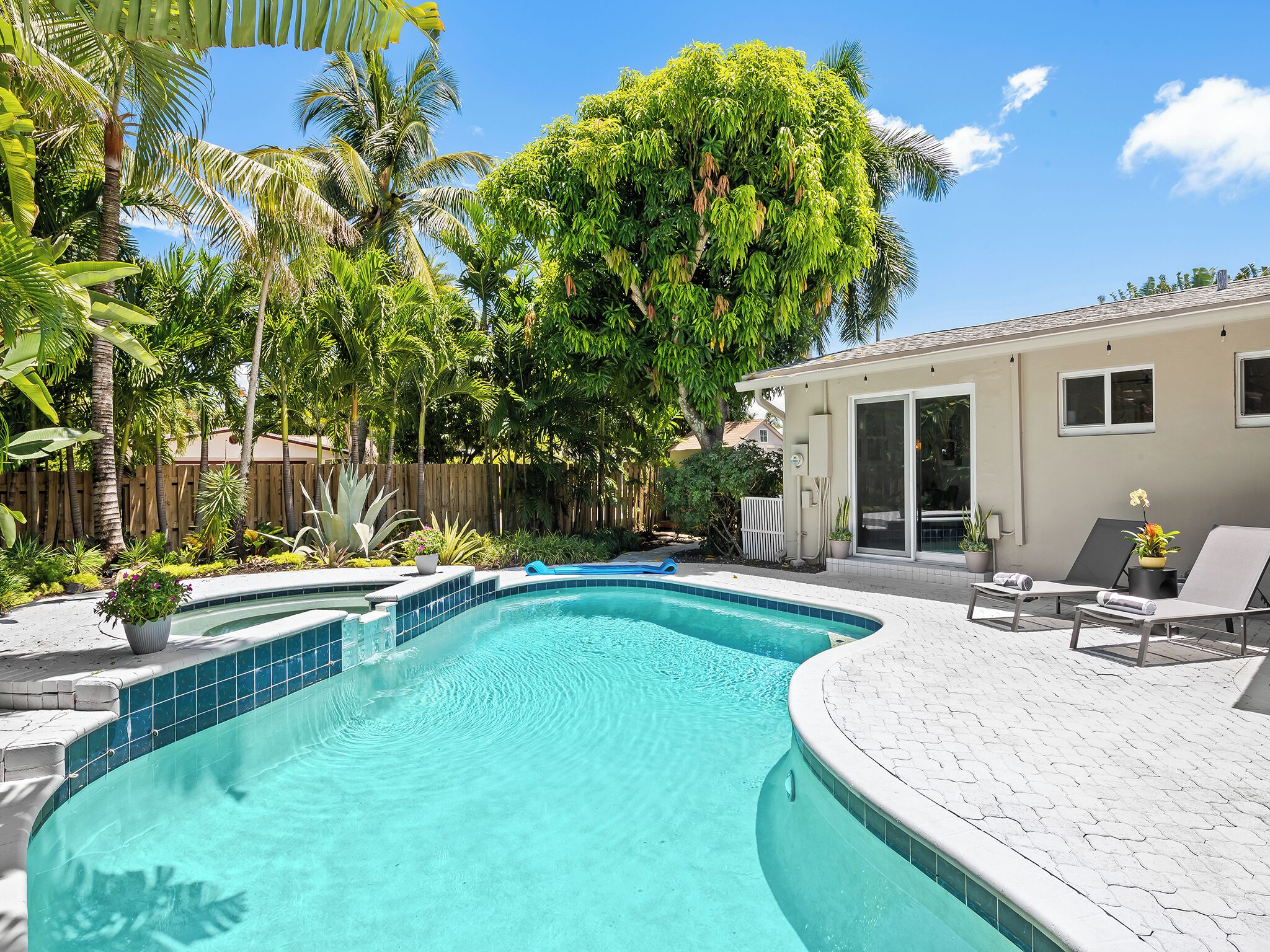 Une piscine dont le bord est revêtu de carrelage, entourée d’une végétation luxuriante et délimitée par une clôture en bois.