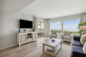 A spacious living room with a flat-screen TV, wooden entertainment unit, and a view of the beach. - Amelia Surf & Racquet Club B150 - 5th Floor Oceanview - Pools - Tennis Courts (Fernandina Beach)