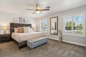 A bedroom with a bed, bedside tables, a ceiling fan, a large window with blinds, and a mirror.