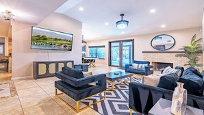 A modern living room with a flat-screen TV, comfortable seating, a fireplace, and a large window offering a view of the outdoors. - New! Private Oasis W Pool, Outdoor BBQ & Gameroom!! (Indio)