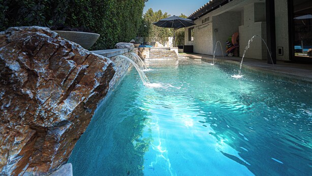 A swimming pool with clear water, a rock feature, and a building with a tiled roof in the background. - New! Private Oasis W Pool, Outdoor BBQ & Gameroom!! (Indio)