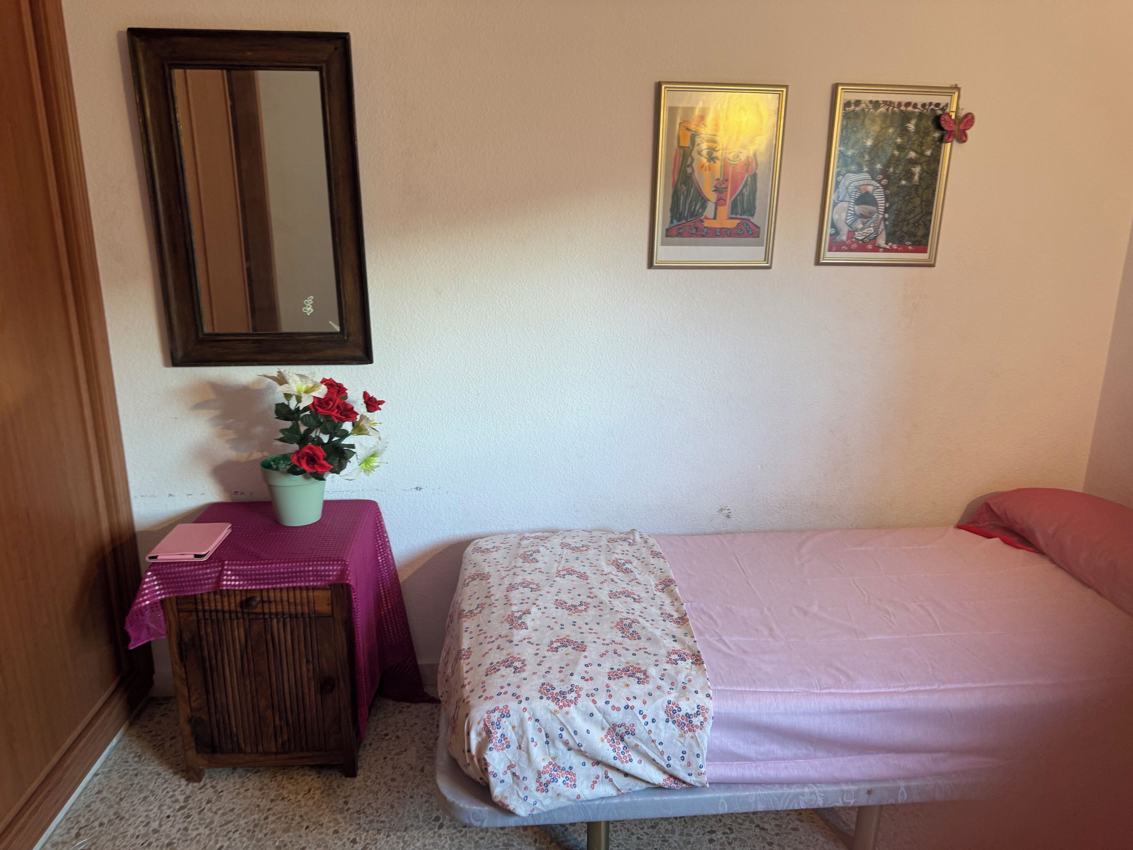 Une petite pièce comprenant un lit, une table de chevet recouverte d’une nappe rose, un miroir et deux tableaux encadrés accrochés au mur.