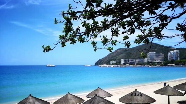 A beach with thatched umbrellas, clear blue water, and buildings in the background. - Sanya Seaside Breeze Homestay (Sanya)