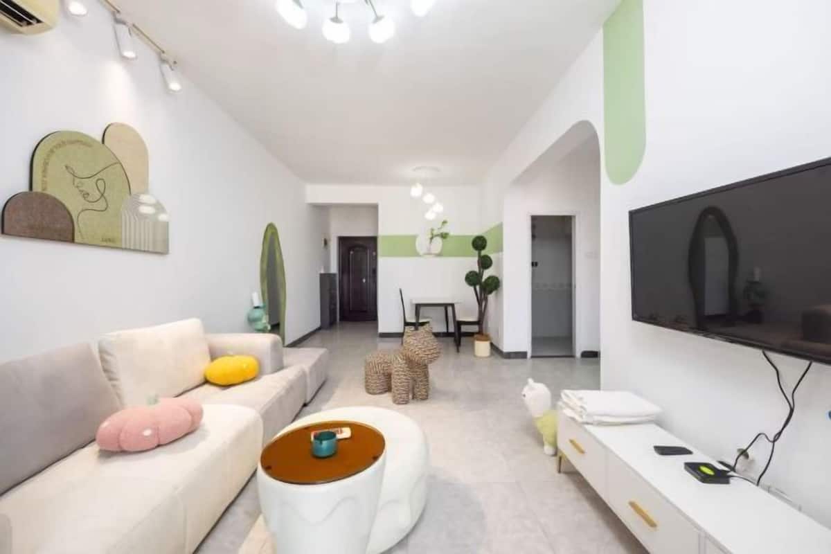 Een moderne woonkamer met een bank, een poef, een flatscreen televisie en een eethoek met stoelen en een tafel.