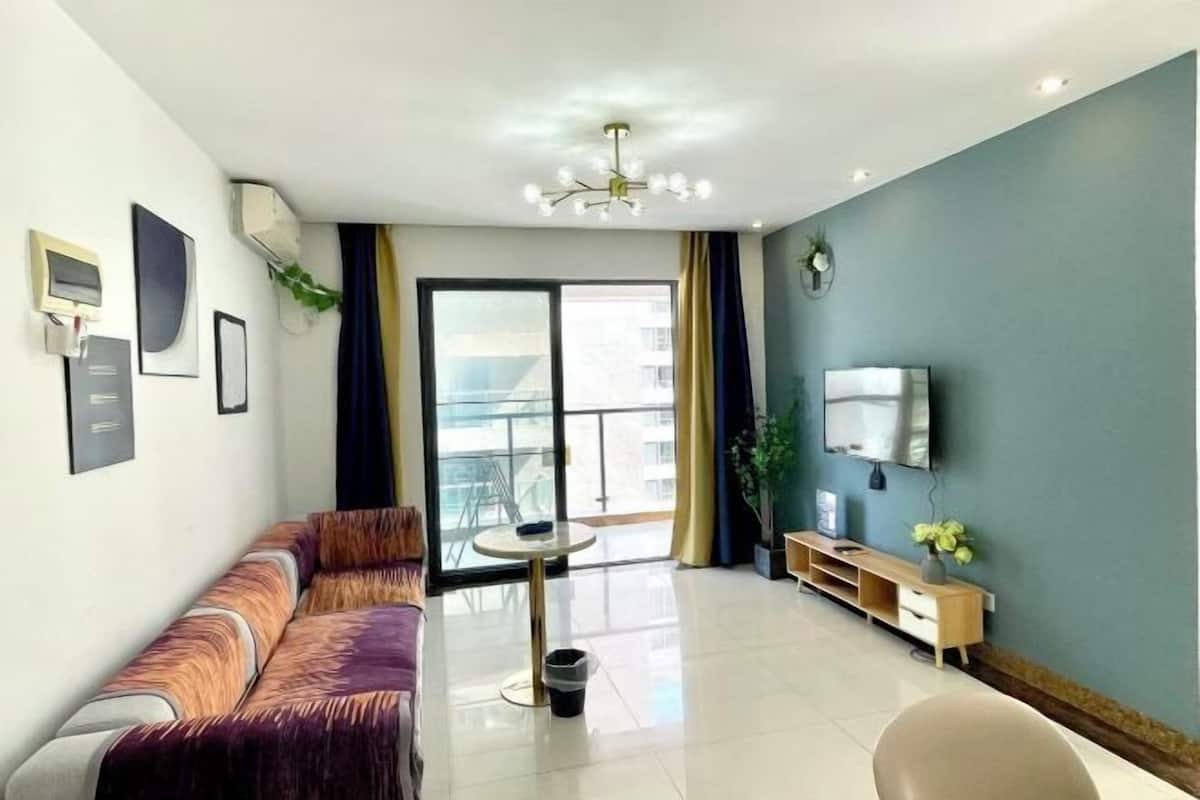 Een moderne woonkamer met een bank, een klein rond tafeltje, een aan de muur bevestigde televisie en een balkon met uitzicht.