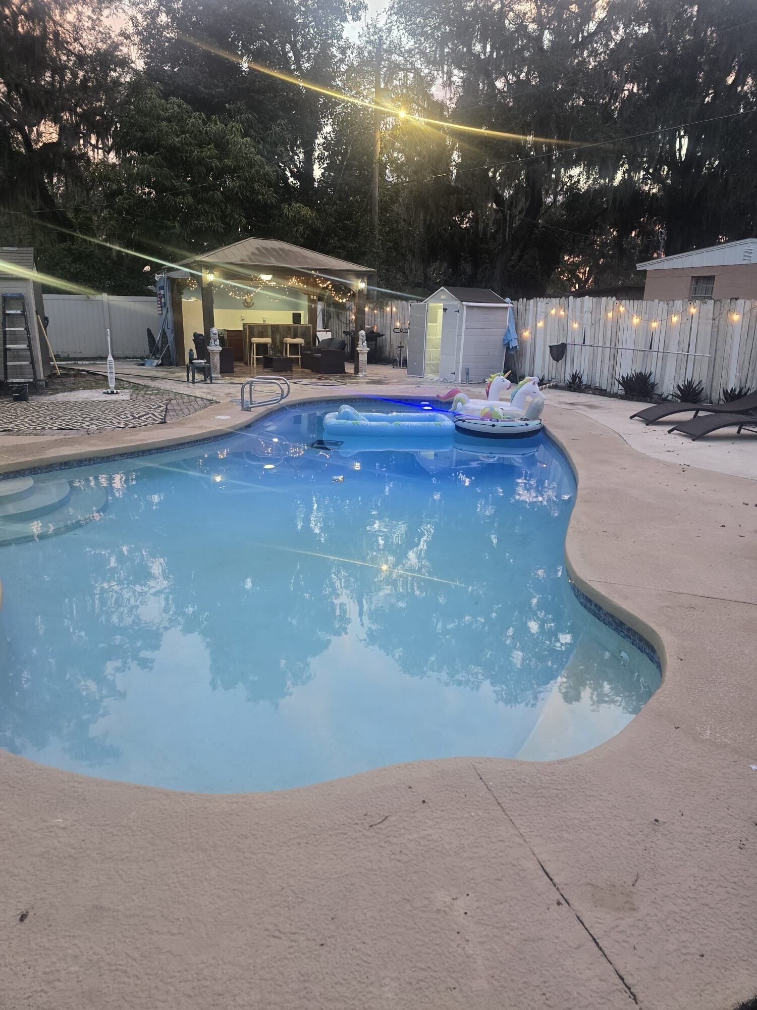 Ein Pool mit geschwungener Kante, umgeben von einer Liegefläche mit Liegestühlen und einem kleinen Gebäude mit Lichterketten.