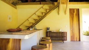 Una escalera de madera que lleva al segundo piso, una zona de cocina con cafetera y una puerta de madera.