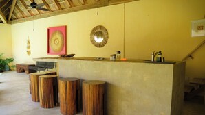 Zona de bar con mostrador de concreto, taburetes de madera, ventilador de techo y decoración en las paredes, incluyendo un espejo y un cuadro enmarcado.