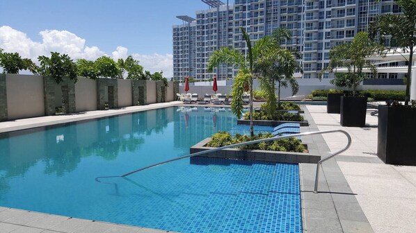 A swimming pool with lounge chairs and umbrellas, surrounded by a low wall and potted plants. - 2 Bedroom Mactan Newtown Beach Condo  (Lapu-Lapu)