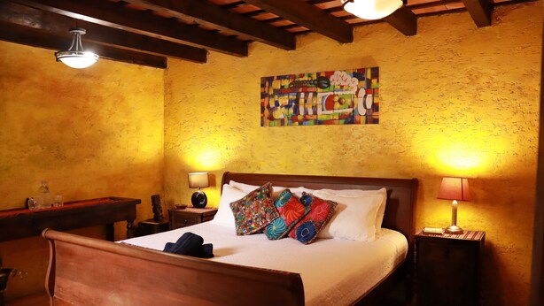 Un dormitorio con cama de madera, dos mesitas de noche con lámparas, un cuadro abstracto colorido en la pared y suelo de baldosas.