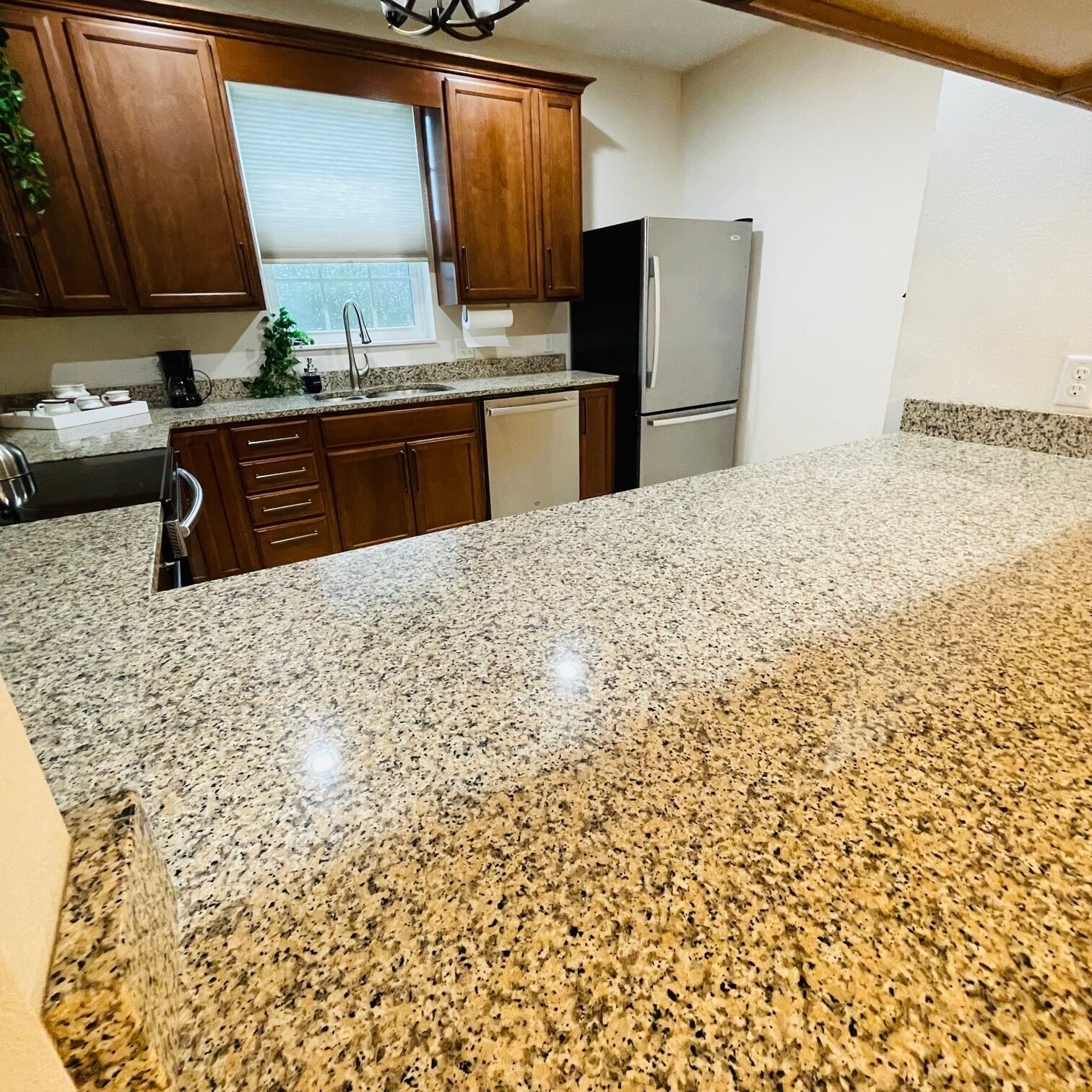 A kitchen with granite countertops, wooden cabinets, a stainless steel refrigerator, and a window with blinds.