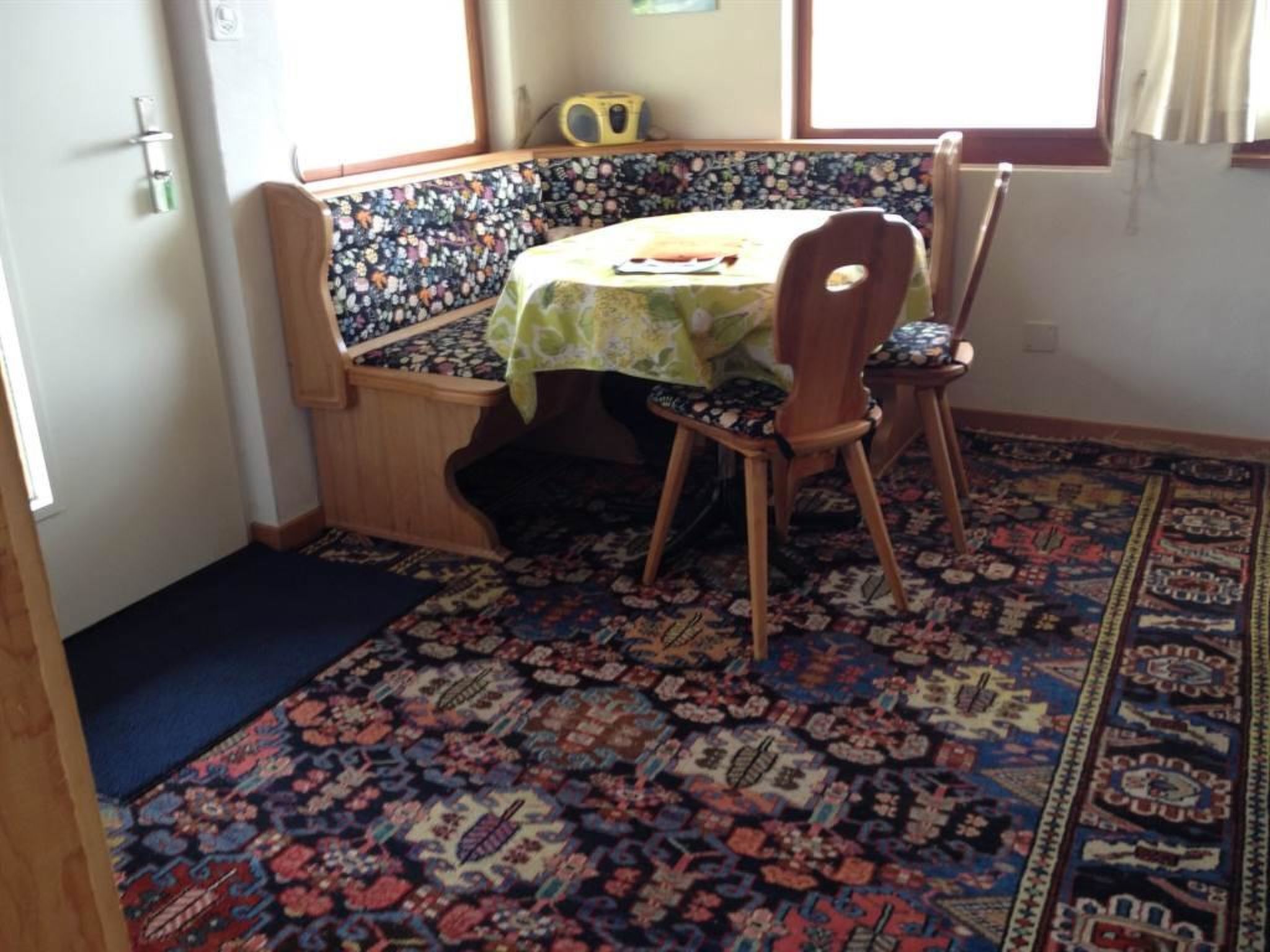 A room with a corner bench, a table with a tablecloth, a patterned rug, and a window with curtains.