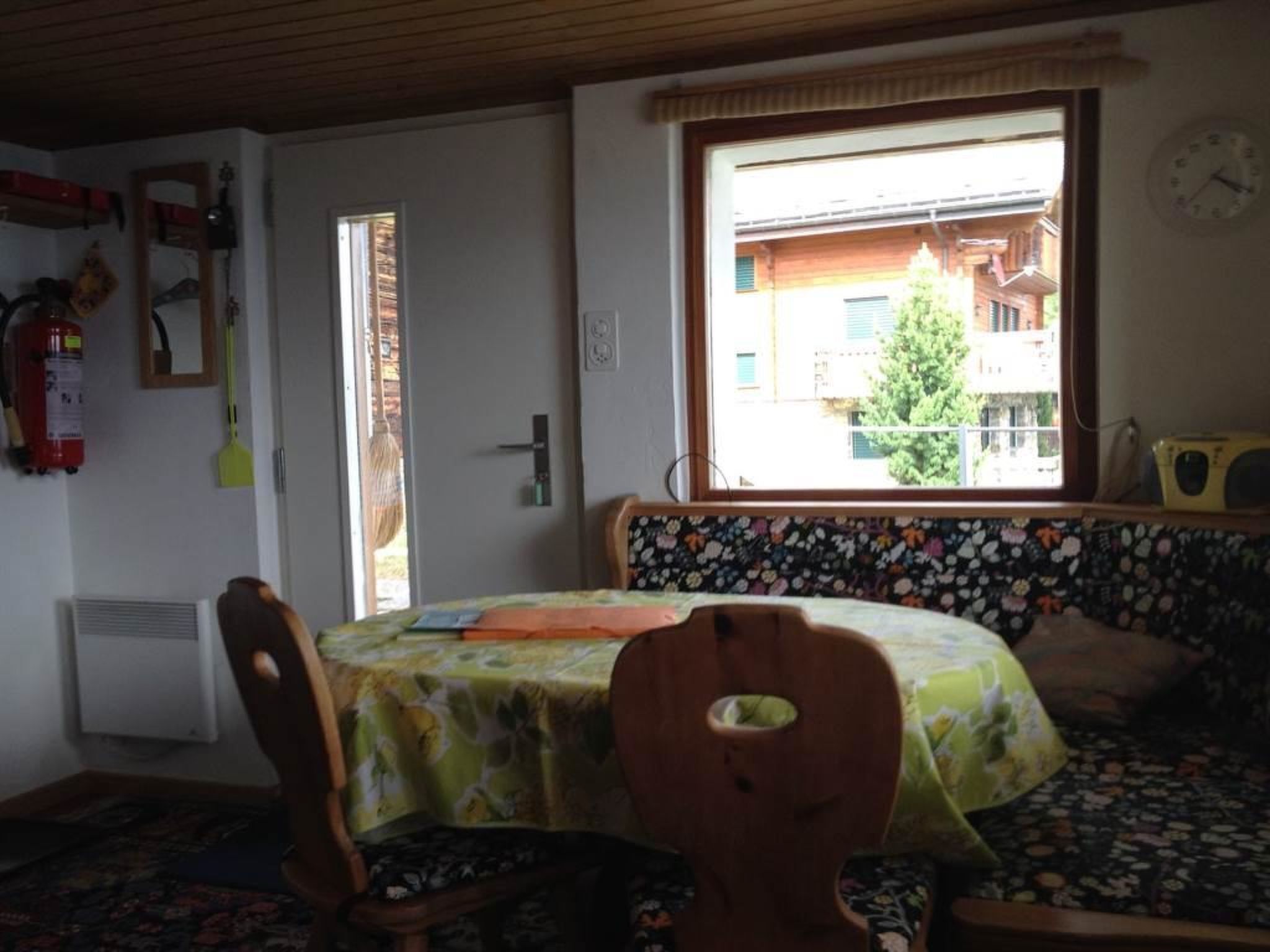 Een eetruimte met een tafel en stoelen, een raam met uitzicht op gebouwen, een brandblusser en een klok aan de muur.