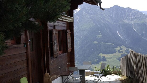 Een houten hut met een stenen terras, twee klapstoelen en uitzicht op bergen.