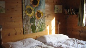 Een kamer met een bed, een wandtapijt met zonnebloemen, een boekenkast en een raam met gordijnen.