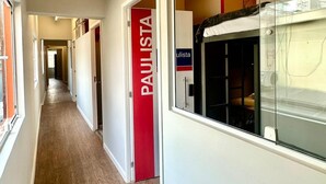 A corridor with a wooden floor, a red door labeled "PAULISTA", and a window with a view of a room with a bed and a red wall. - Hostel Distrito 011 (São Paulo)