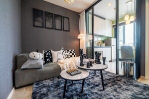Ein modernes Wohnzimmer mit einem grauen Sofa, einem Couchtisch und einem Teppich. An den Wänden befinden sich gerahmte Bilder und ein Fenster mit Vorhängen.