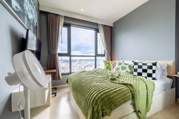 Ein modernes Schlafzimmer mit großem Fenster, einem Bett mit grüner Bettwäsche, einem Standventilator und einem an der Wand befestigten Fernseher.