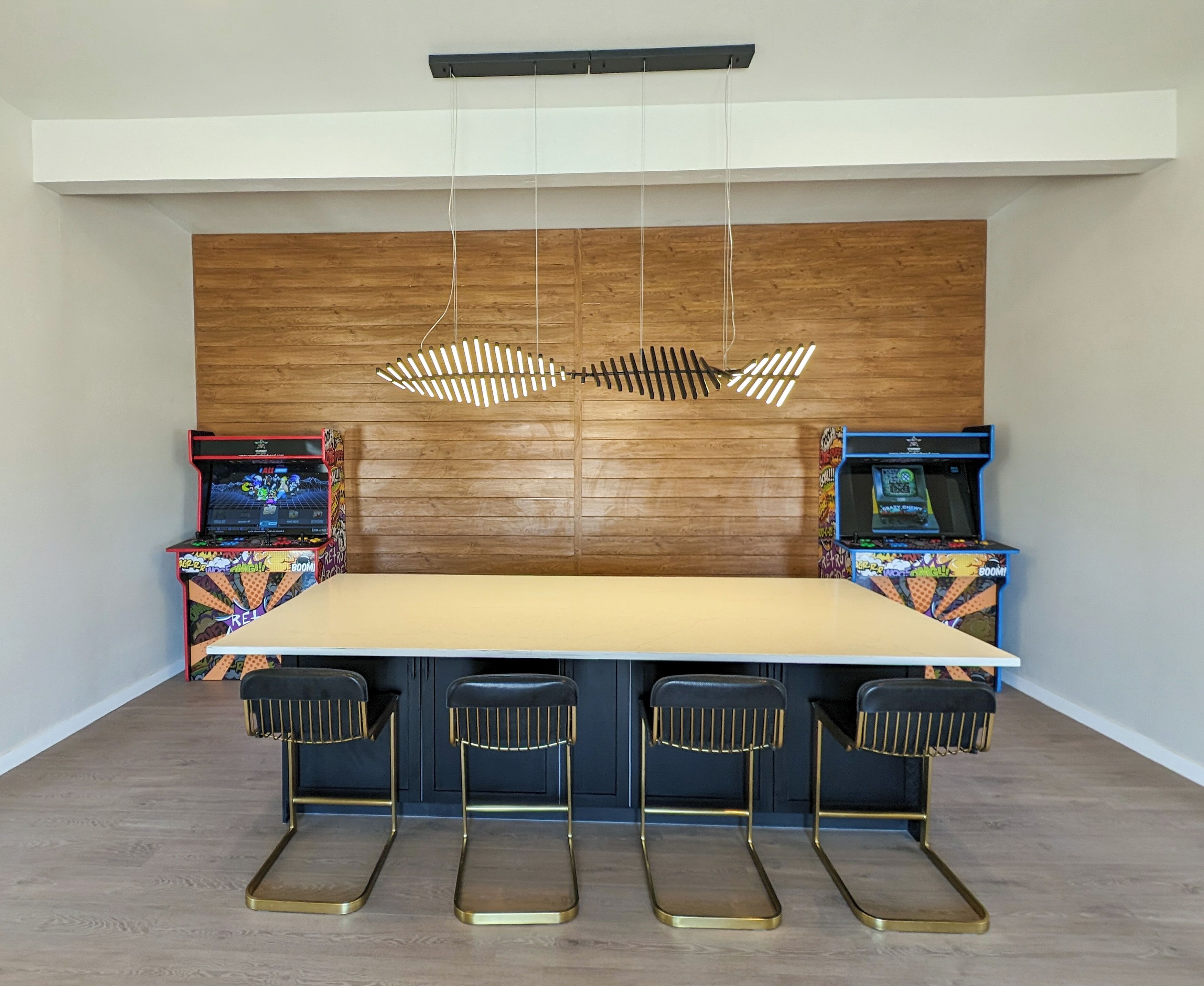 A modern dining area with a long table, four bar stools, and two arcade machines.