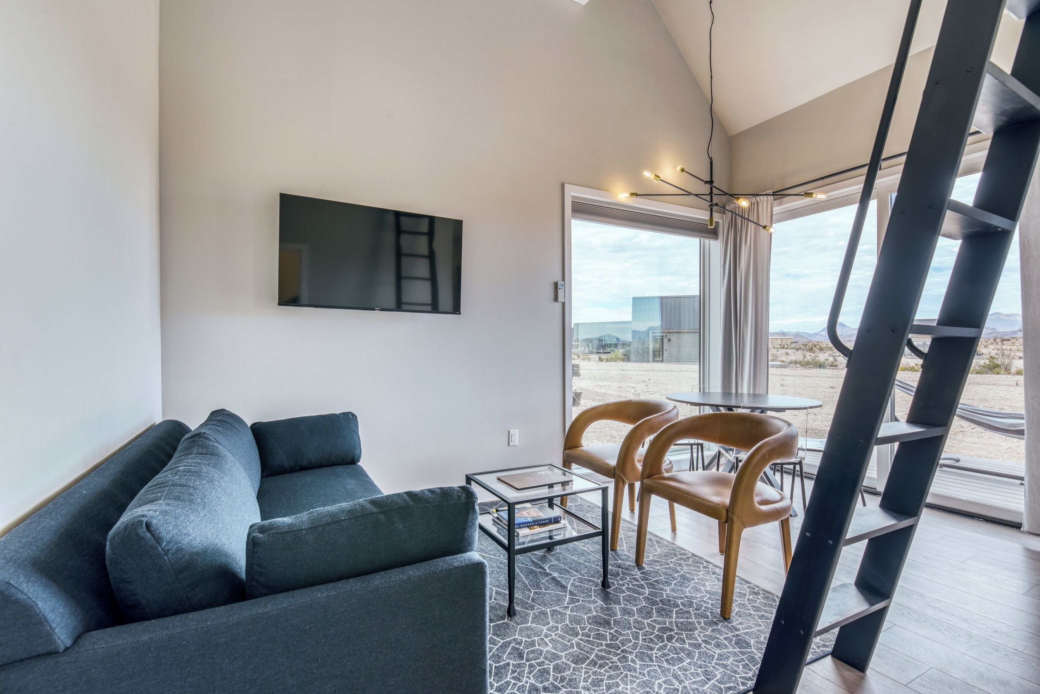 Ein modernes Wohnzimmer mit einer Couch, einem Couchtisch und einem an der Wand befestigten Fernseher. Große Fenster bieten einen Ausblick nach draußen.