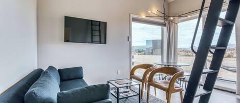 Ein modernes Wohnzimmer mit einer Couch, einem Couchtisch und einem an der Wand befestigten Fernseher. Große Fenster bieten einen Ausblick nach draußen.