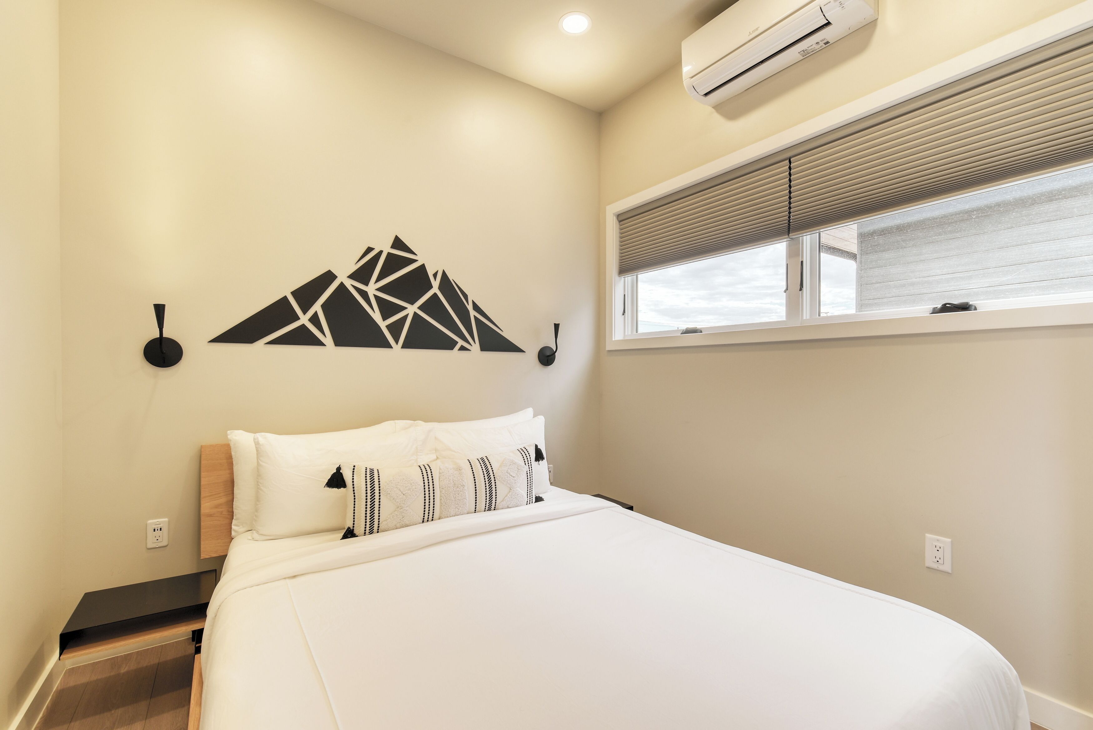 A neatly made bed with white bedding, a geometric wall decoration, a window with blinds, and an air conditioning unit.