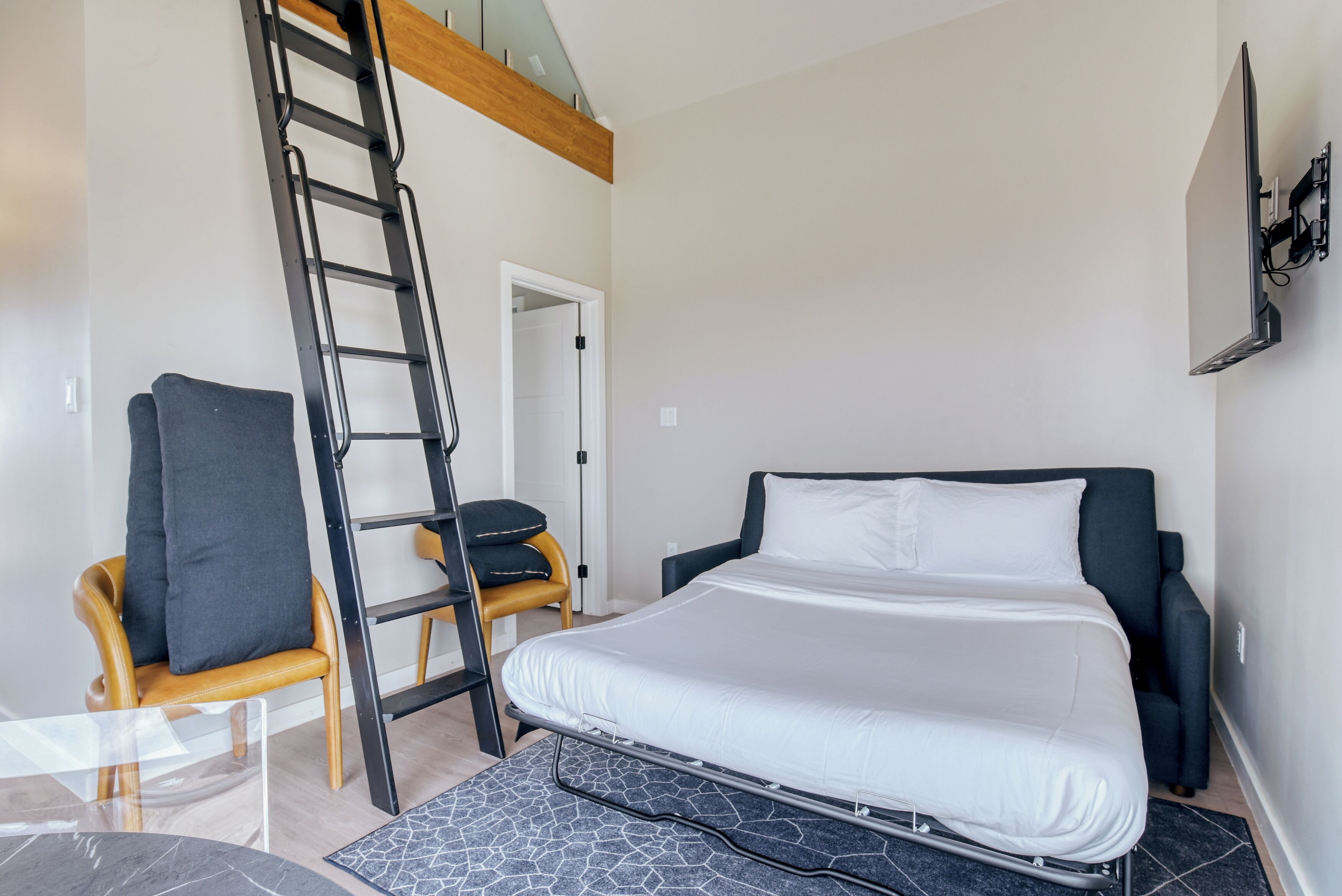 A bedroom with a bed, a chair, a ladder, and a TV mounted on the wall.