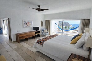 A bedroom with a bed, a TV, a desk, a chair, and a view of the ocean.