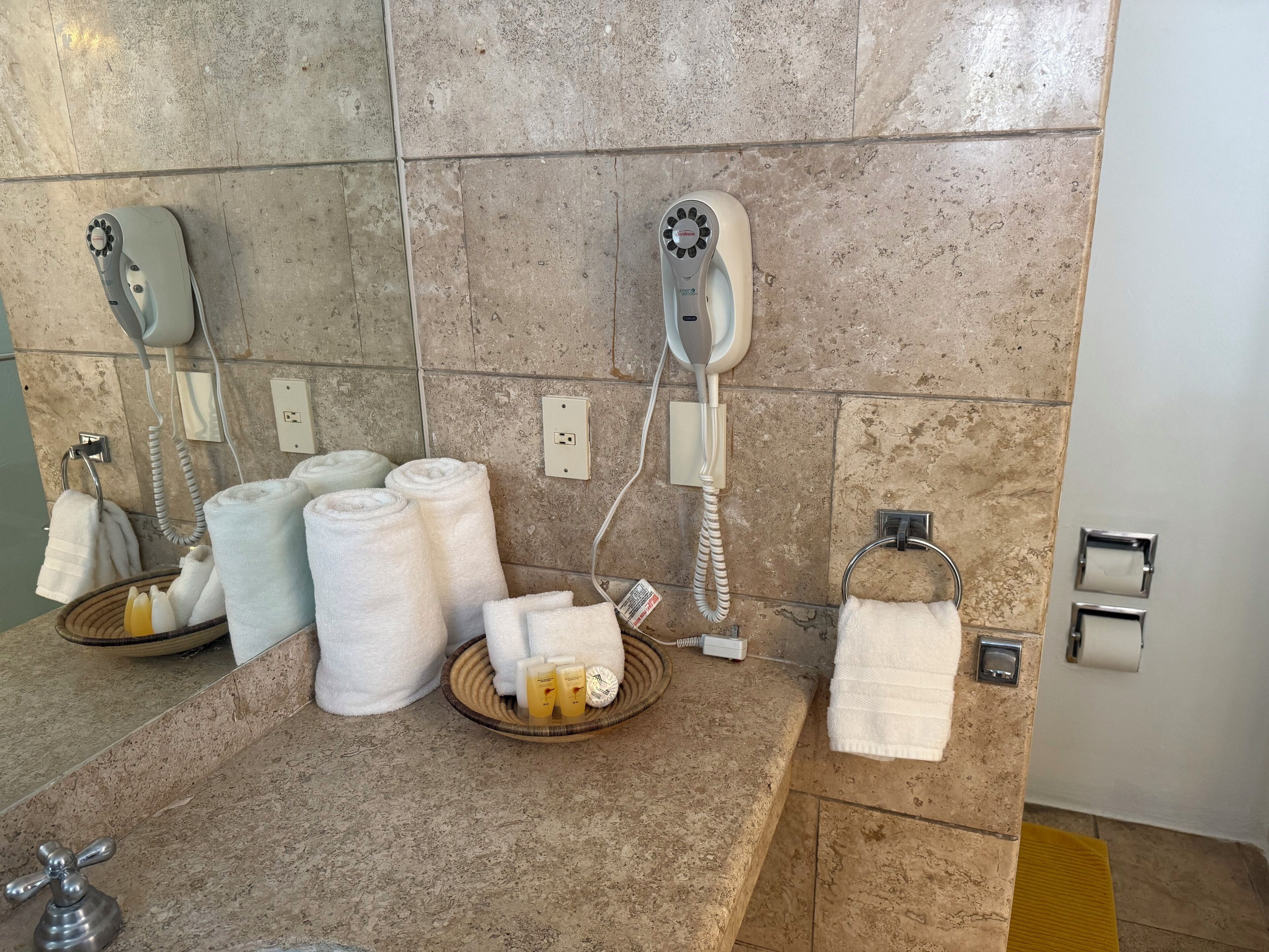 A bathroom with two hair dryers, towels, toiletries, and a hand dryer.