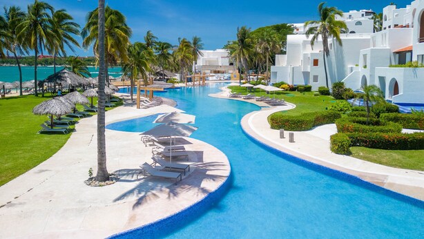 Une piscine d’un complexe hôtelier, bordée de palmiers, avec des chaises longues et des bâtiments blancs.