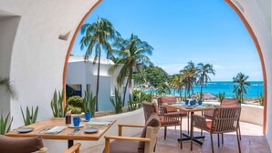 Une terrasse avec des tables et des chaises, donnant sur une plage bordée de palmiers et sur l’océan.