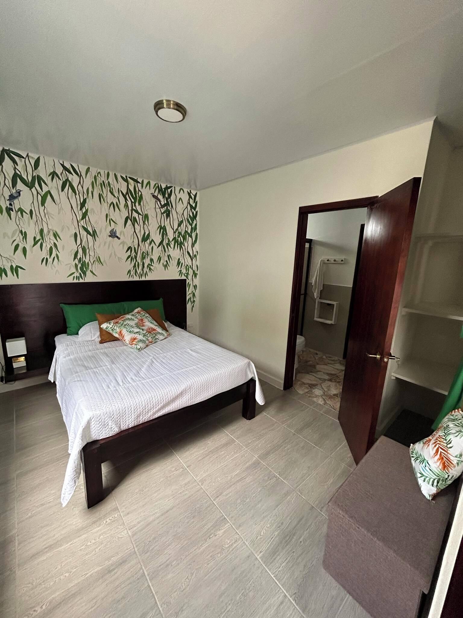 Una camera da letto con un letto, un comodino, una parete con motivo a foglia e una porta che conduce in un'altra stanza.