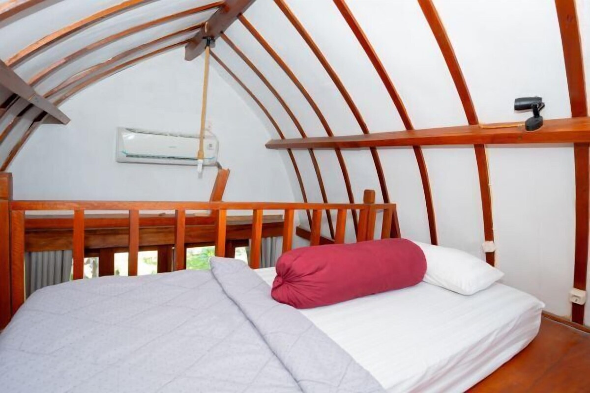 A single bed with a wooden headboard, a red pillow, and a white pillow in a room with a curved wooden ceiling and an air conditioning unit on the wall.
