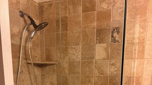 A glass-enclosed shower stall with a handheld showerhead, a wall-mounted shelf, and a handheld shower control.