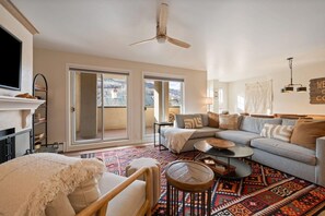 Een ruime woonkamer met een bank, fauteuils, een salontafel en een open haard. Er zijn grote ramen en een plafondventilator.