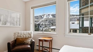 Una habitación acogedora con un ventanal grande que ofrece vistas de una montaña nevada, un sillón de cuero con un plaid estampado y una pequeña mesa de madera.