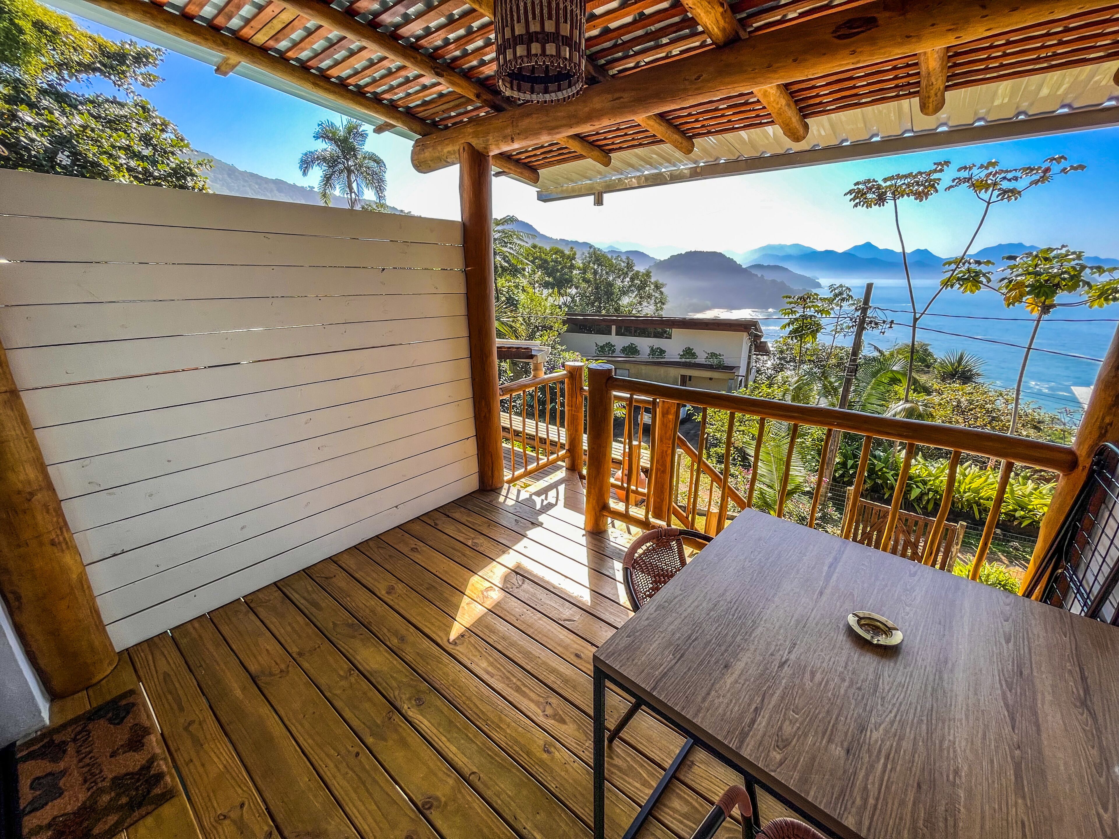 A wooden deck with a table and chairs, overlooking a mountainous landscape with a view of the sea.
