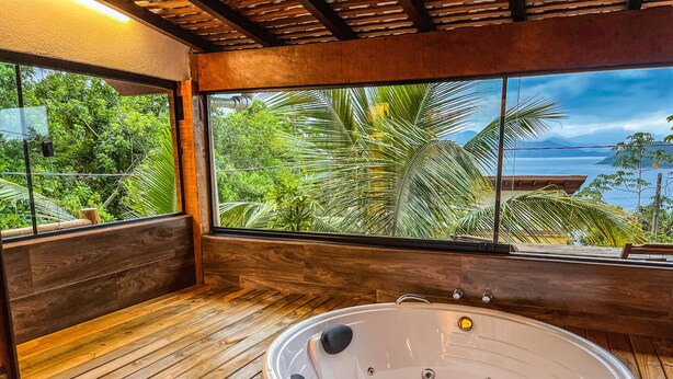 A wooden spa area with a hot tub, wooden benches, and a large window offering a view of lush greenery and a distant mountain. - Casa Varandas (Ubatuba)